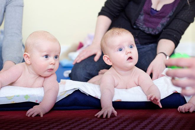 Foto: Zwei Babys liegen auf dem Bauch auf einer Wickelunterlage. Sie schauen das grüne Spielzeug in der Hand eines Erwachsenen mit großen Augen an.