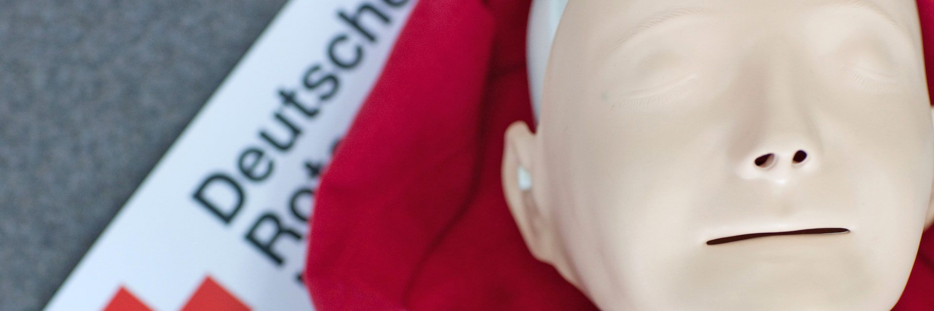 Foto: Eine Übungspuppe liegt auf einer roten Decke. Daneben liegt ein Banner des Deutschen Roten Kreuzes.