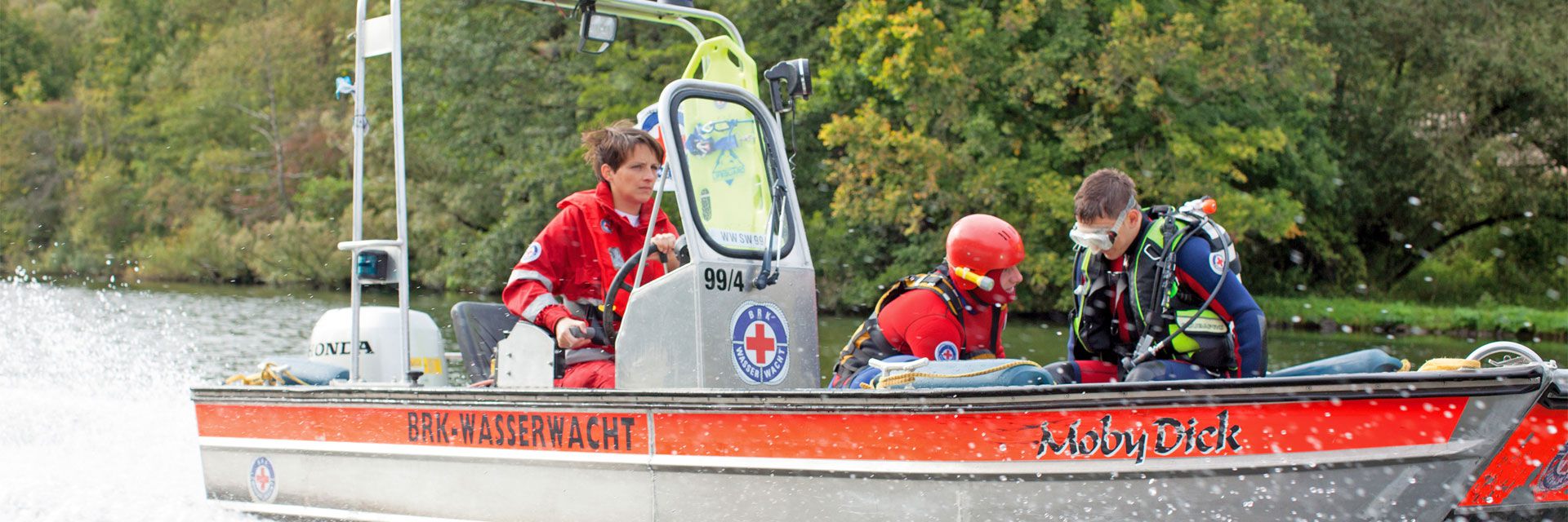 Foto: Zu sehen ist ein Boot der BRK-Wasserwacht in schneller Fahrt. Im vorderen Teil des Bootes sitzen zwei Rettungskräfte. Einer von beiden trägt eine Tacherausrüstung.